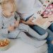 Mother reading a book to her daughter in a cozy bedroom setting with snacks nearby.
