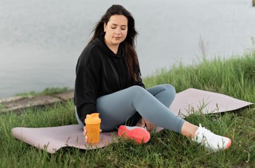 Woman enjoying a peaceful moment by the lake with a yoga mat and water bottle, promoting wellness.